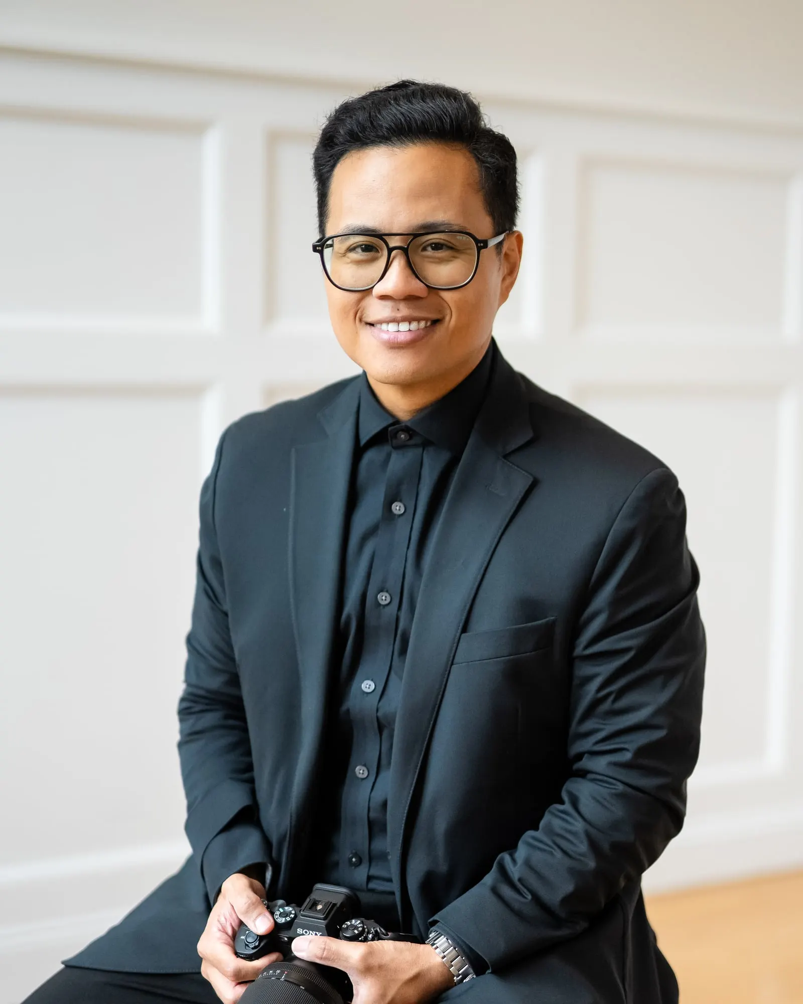 Angelito Jusay, New York City wedding and event photographer, smiling in a dark suit with his camera