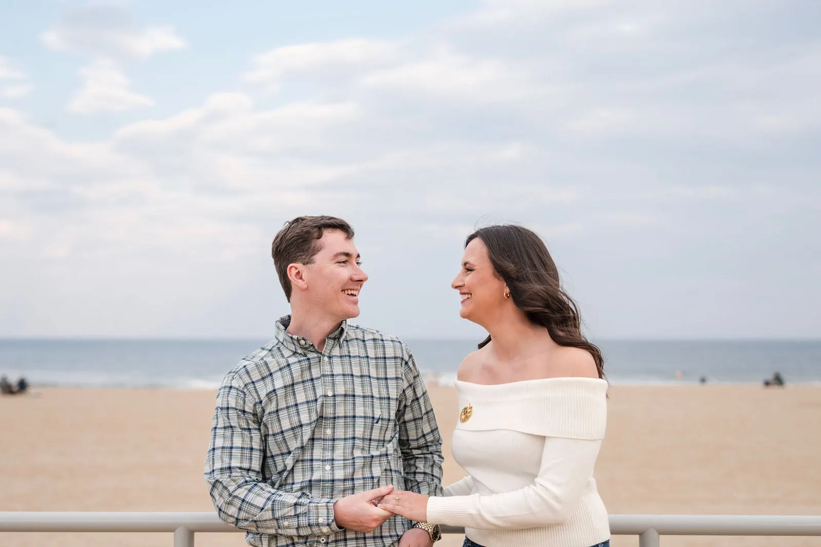 Engaged couple holding hands at the boardwalk railing with the beach and ocean behind them