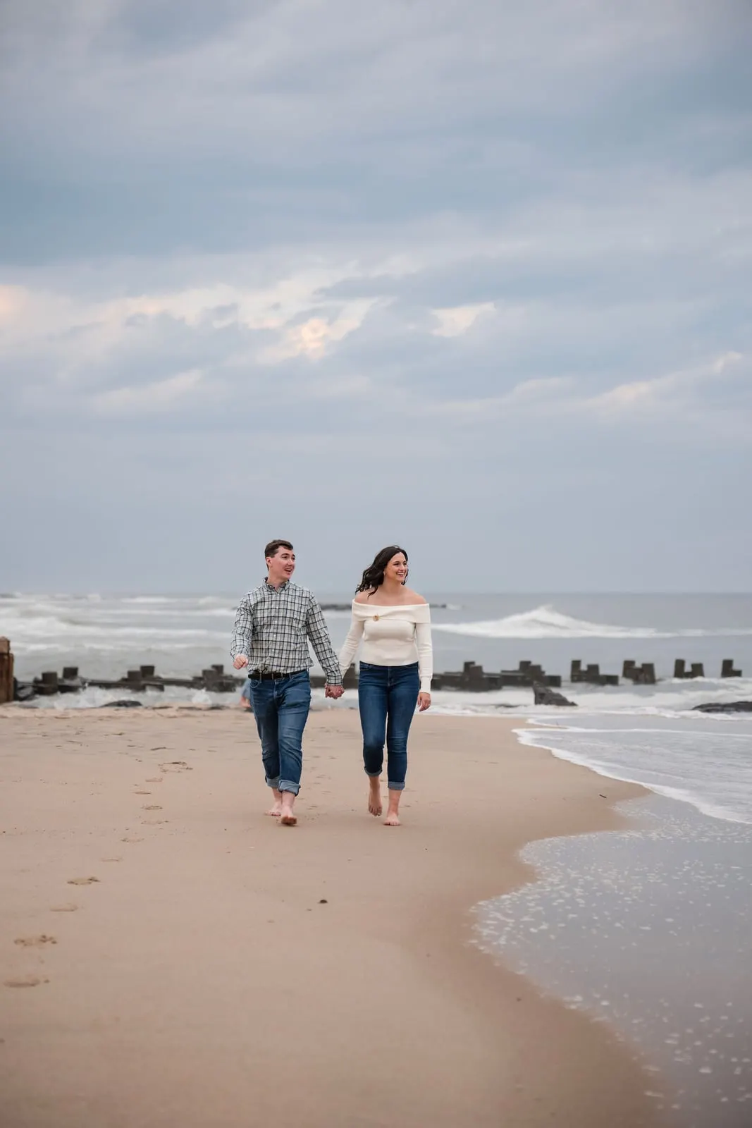 Barefoot engaged couple walking along the shoreline past old wooden pilings under a cloudy sky