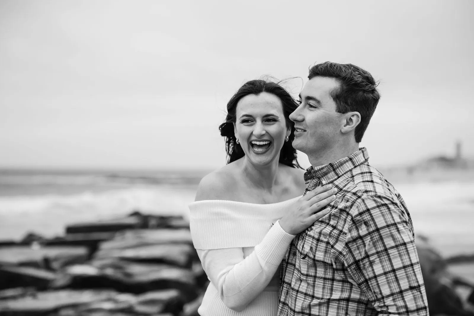Black and white photo of the couple laughing beside a rock jetty