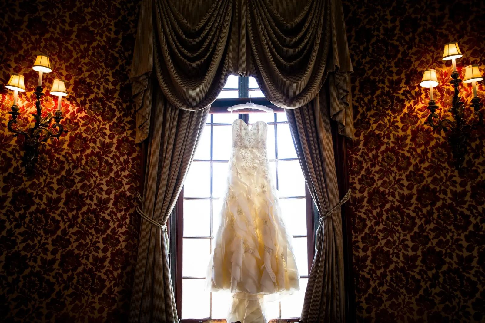 Ruffled wedding gown hanging in a tall window between damask walls and sconces