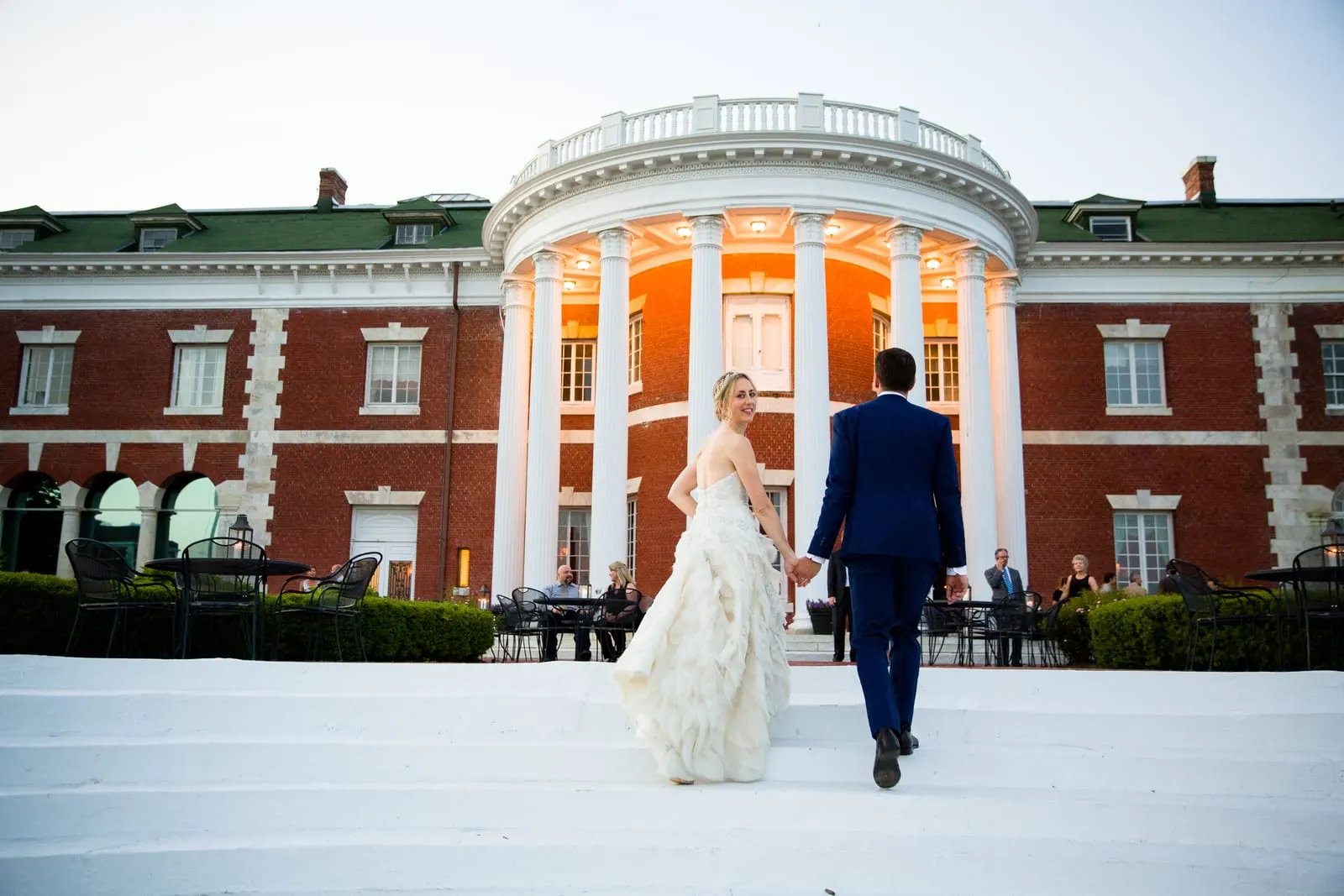 Bride and groom walking in front of Bourne Mansion's glowing columns at sunset