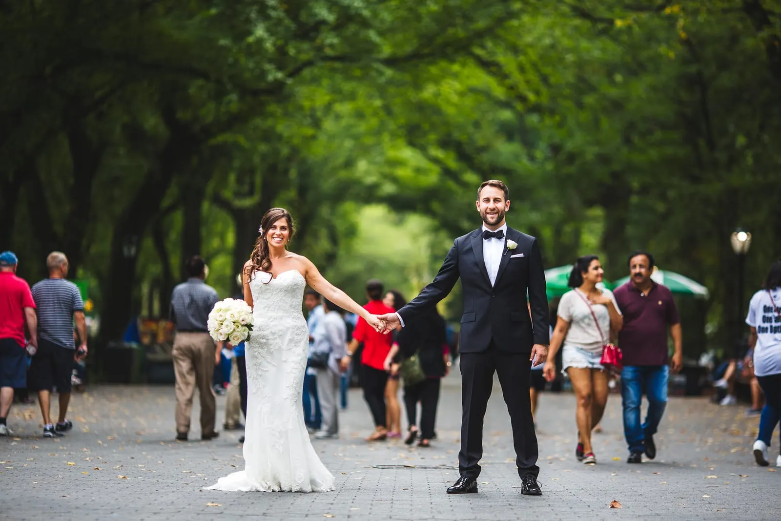 Bride and groom holding hands as crowds stroll the Mall in Central Park