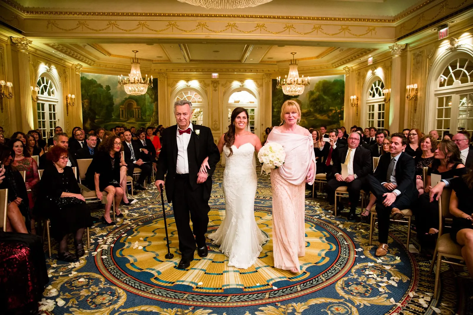 Bride walking down the aisle with her parents under crystal chandeliers