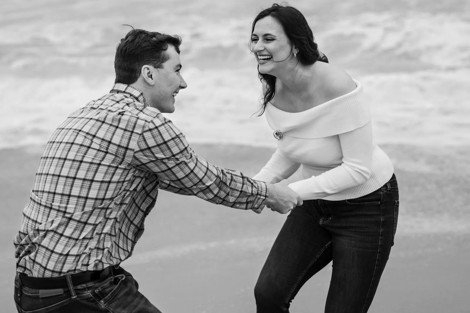 Black and white photo of an engaged couple laughing as they run from a wave on the beach in Asbury Park