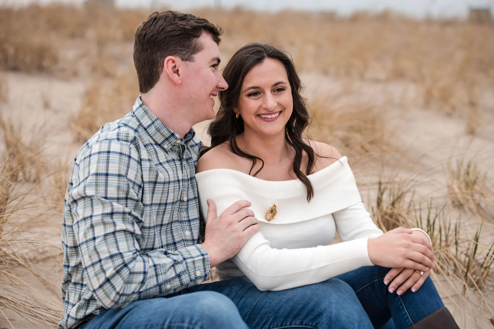 Smiling engaged couple sitting together among golden dune grass on the beach in Asbury Park, New Jersey