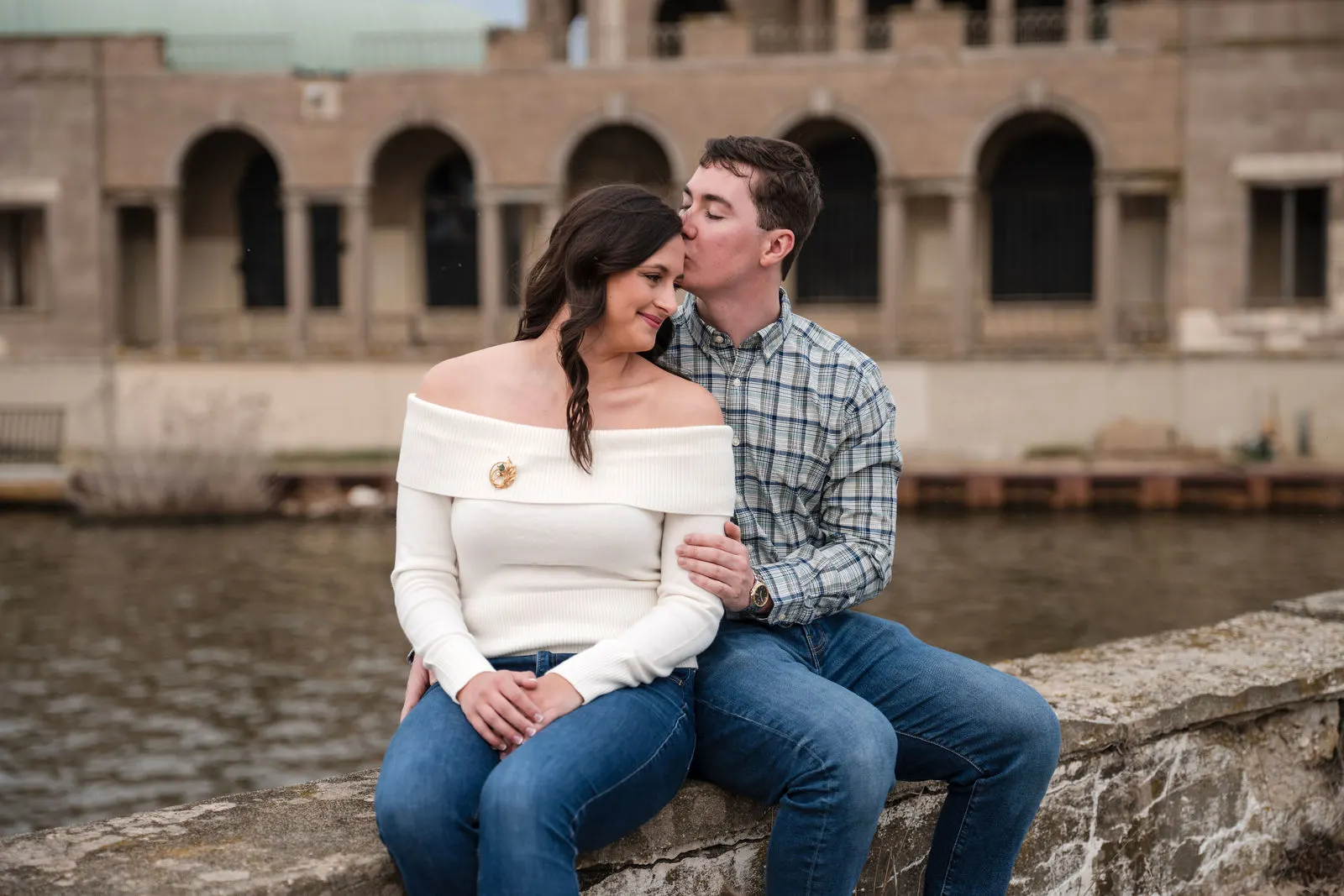 Man kissing his fiancee's forehead as they sit on a stone wall along Wesley Lake in Asbury Park, NJ