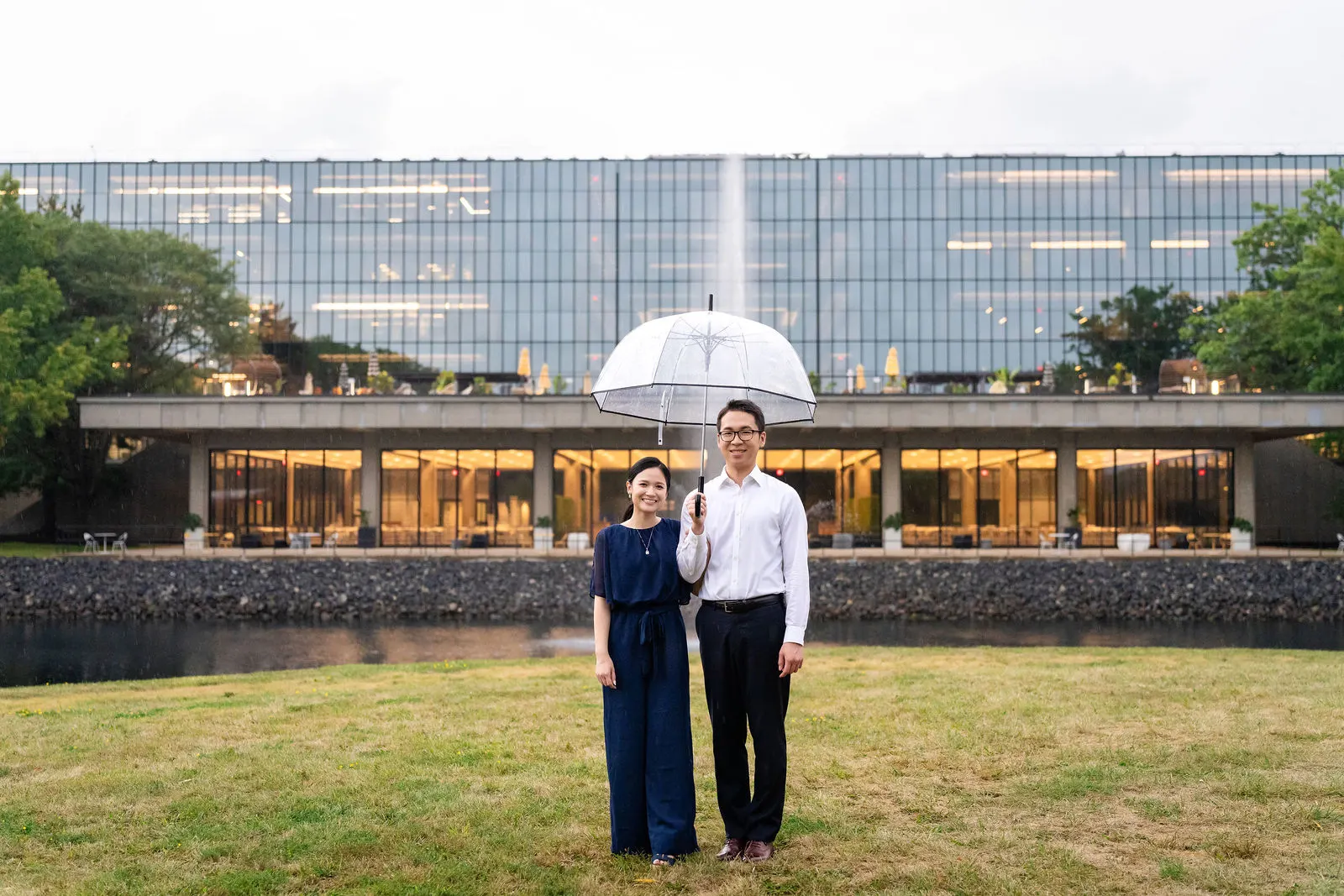 Engaged couple under a clear umbrella on the lawn in front of the Eero Saarinen designed Bell Labs building in Holmdel, New Jersey
