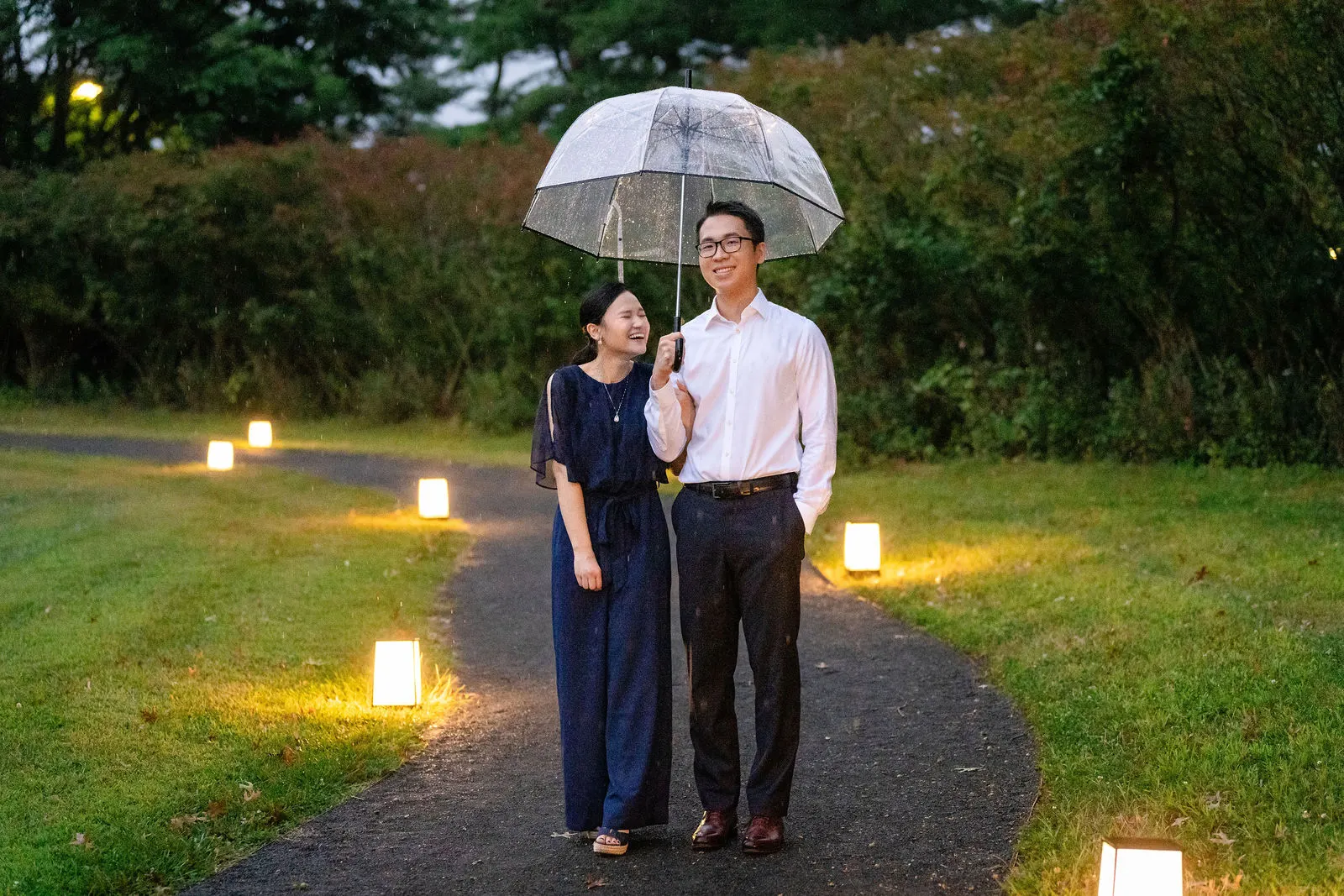 Couple sharing a clear umbrella on a garden path lined with glowing lanterns at dusk in Holmdel, New Jersey