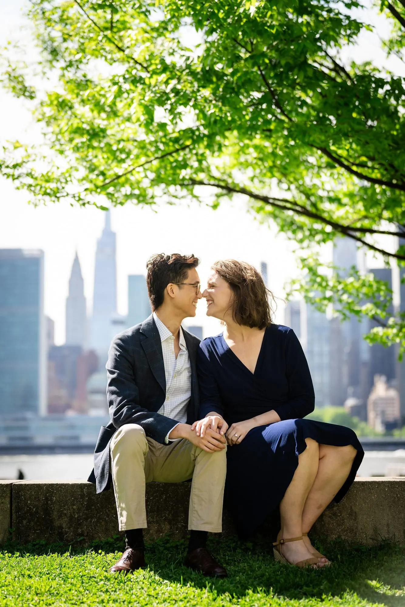 Couple sitting close under the trees with the Manhattan skyline across the East River
