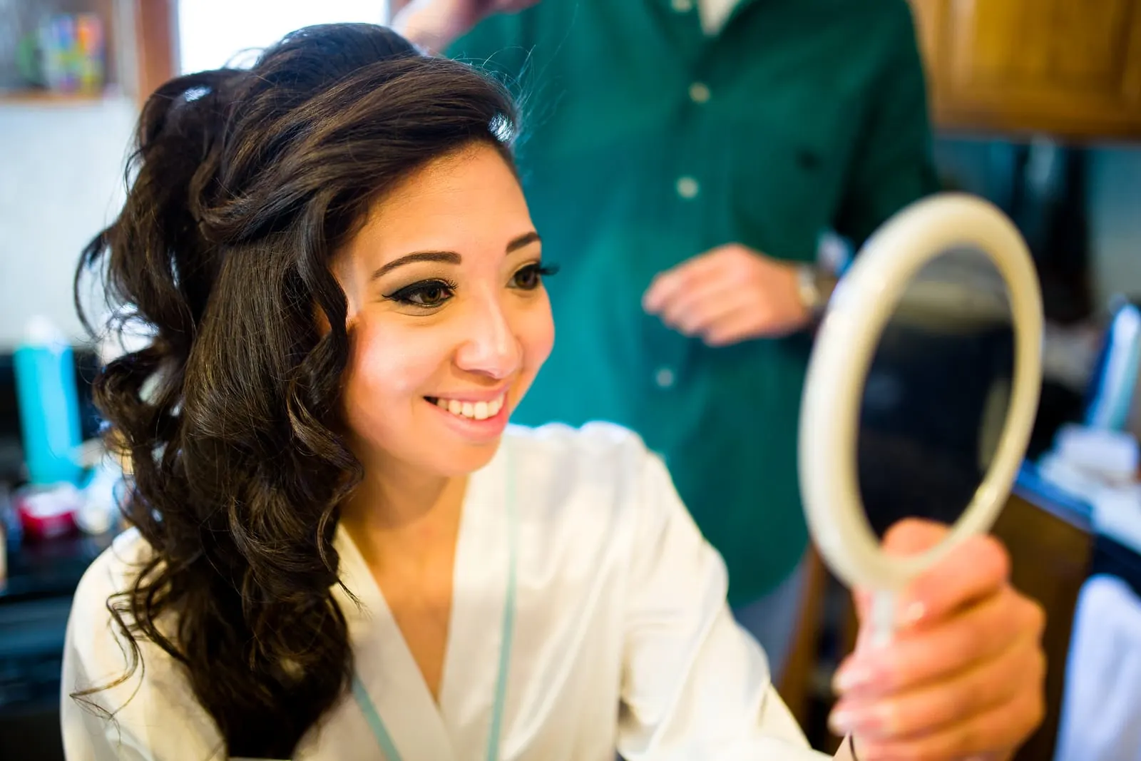 Bride smiling into a hand mirror as her hair is finished