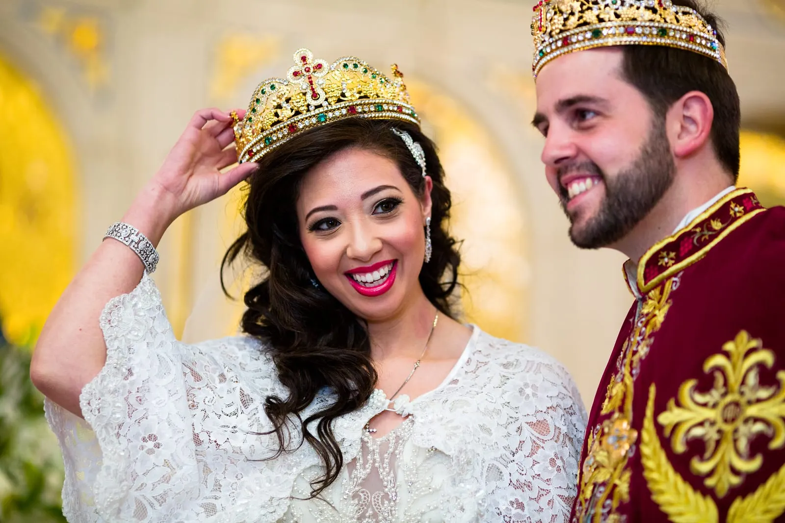 Bride laughing as she adjusts her crown beside the groom in his red and gold cape