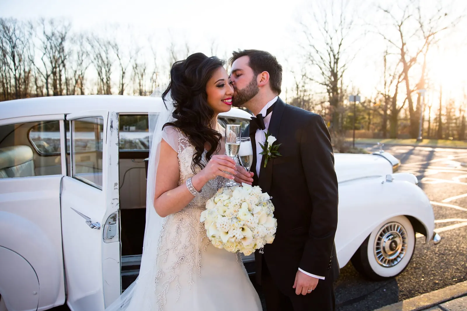 Newlyweds kissing beside the vintage white car with champagne in hand
