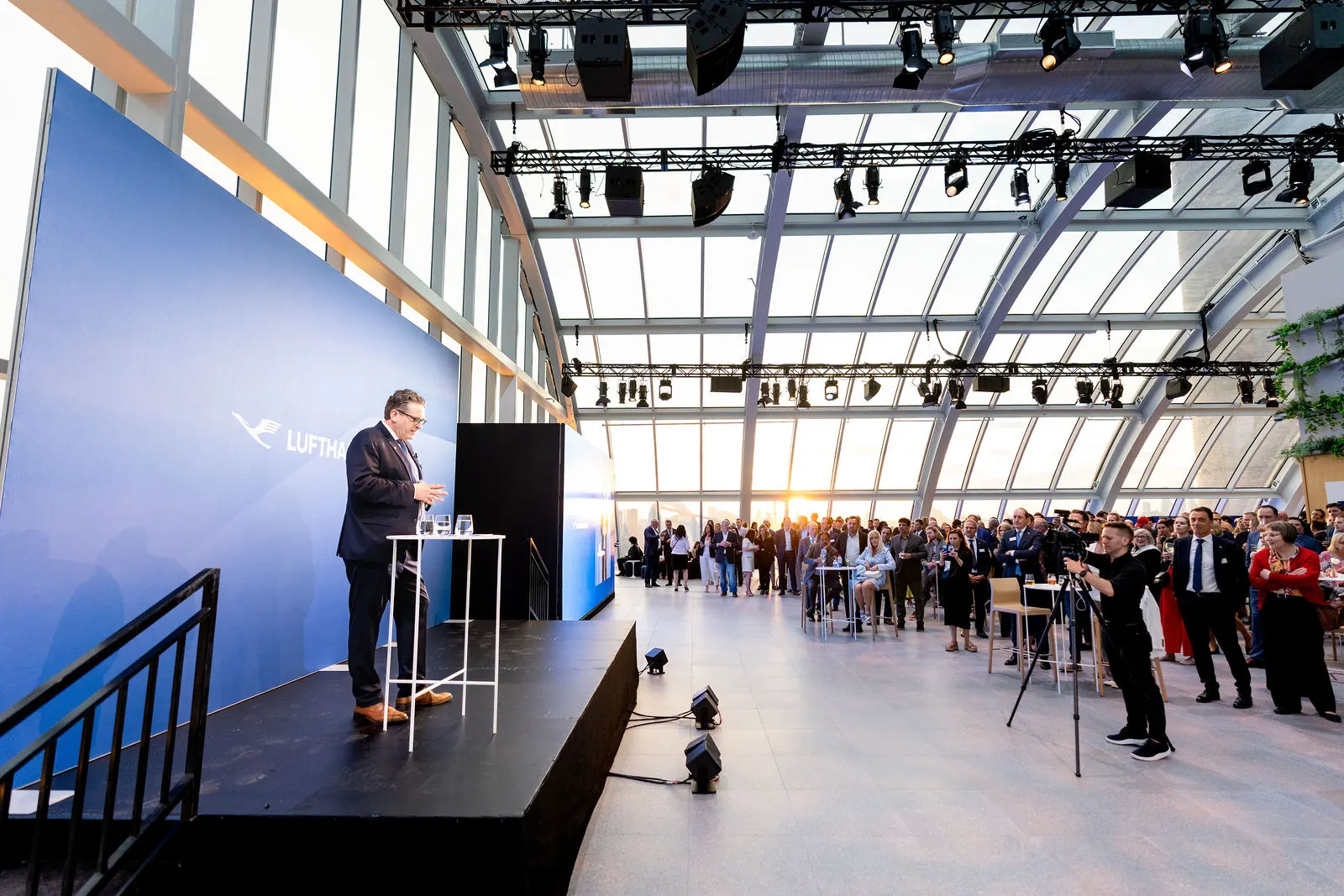 Keynote speaker on a Lufthansa-branded stage addressing a crowd under a glass roof at sunset, Lufthansa 100th anniversary in New York