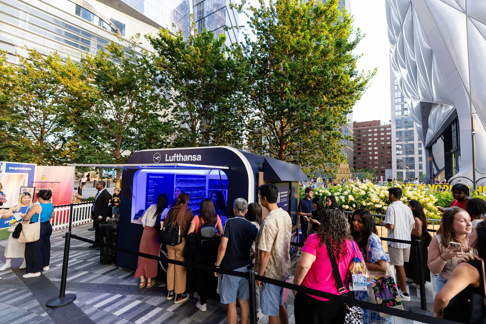 Glowing Lufthansa booth with a long line of guests at golden hour, The Shed and Hudson Yards towers behind