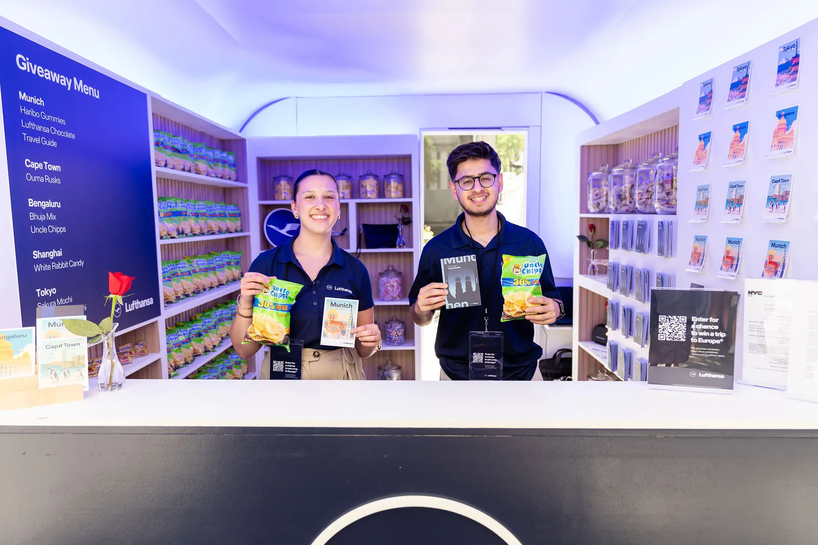 Two smiling Lufthansa staff holding destination treats and travel guides inside the booth at Bryant Park