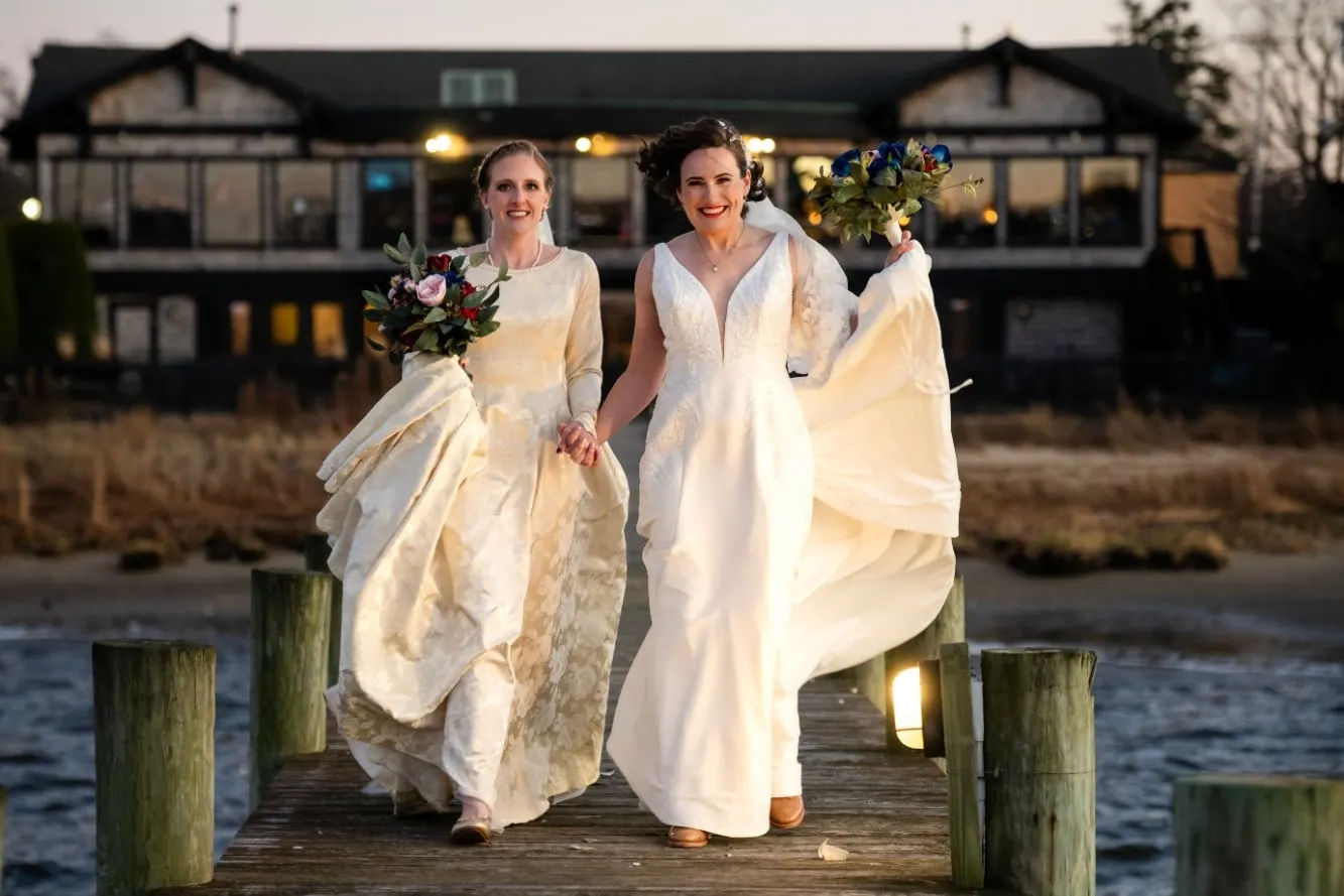 Two brides walking hand in hand down a wooden dock at sunset with the Salt Creek Grille behind them in Rumson, New Jersey