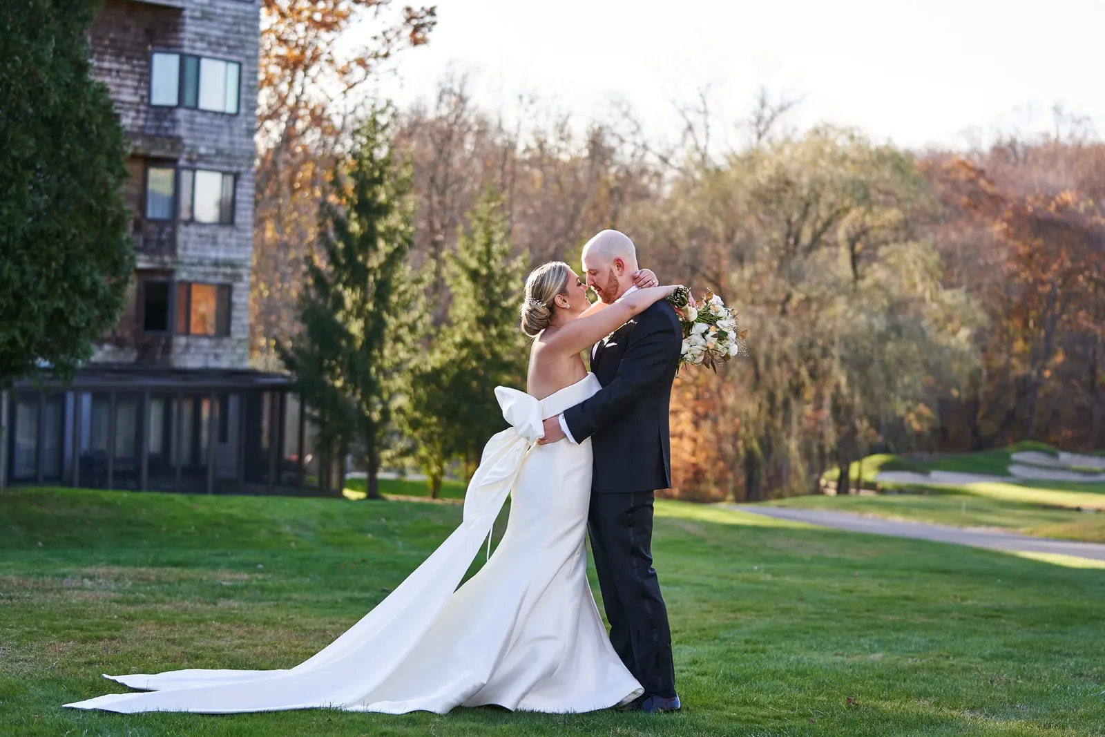 Bride and groom embracing on the lawn with fall foliage behind them