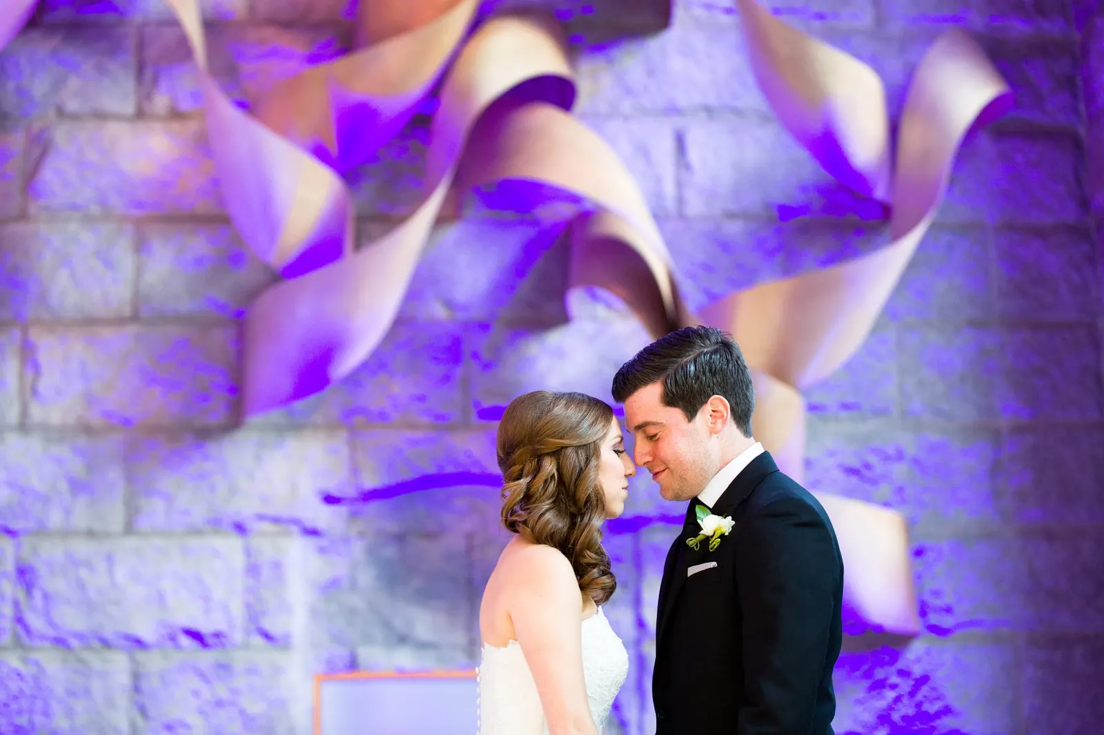 Bride and groom sharing a quiet moment in front of a sculptural wall in violet light